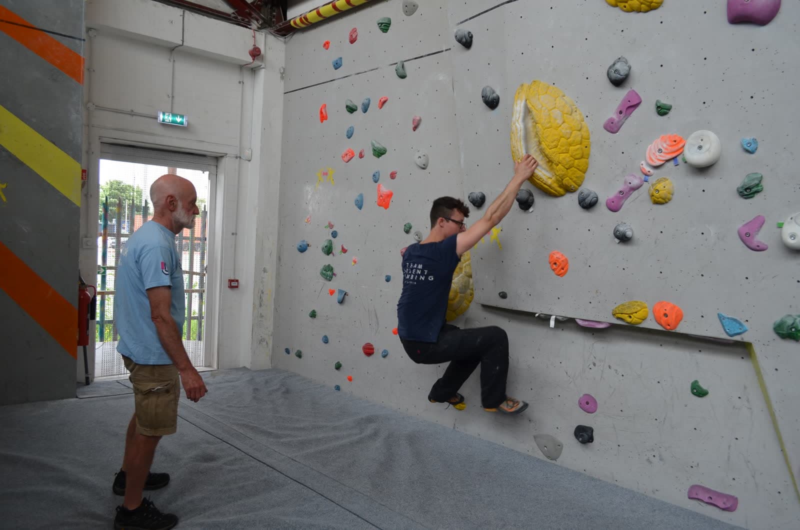 A climber on the wall with a coach watching from the mats