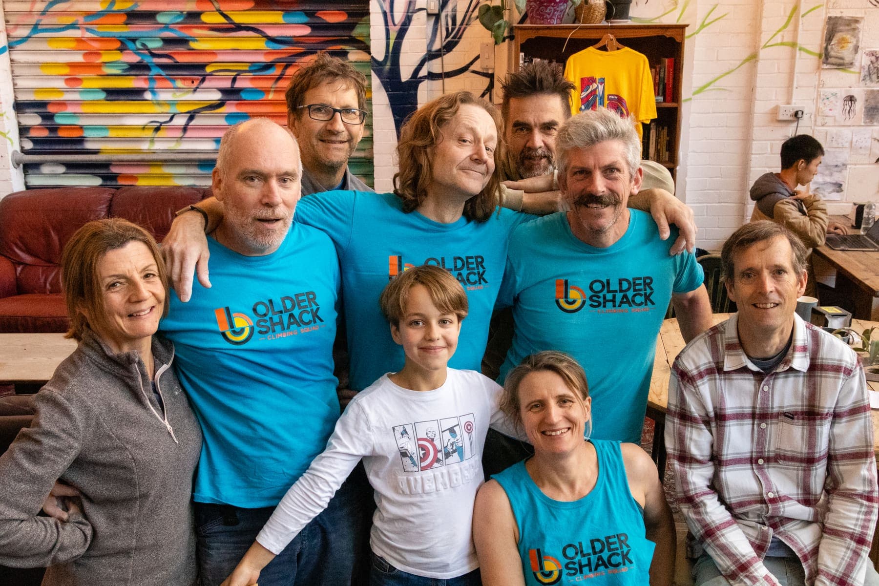 Eight climbers of all ages smiling for the camera in the café, most of them in blue Older Shack T-shirts