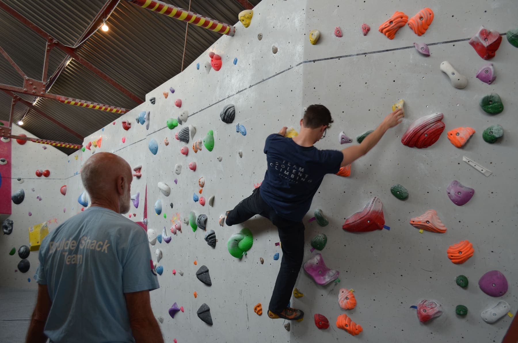 A climber reaching across the wall while a coach watches from the mats