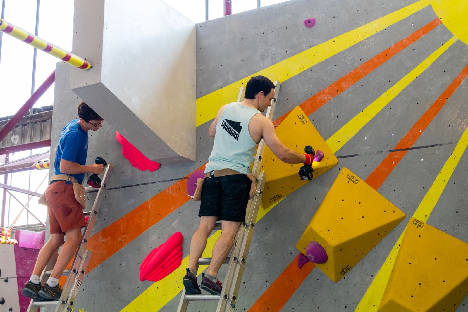 Two route setters on ladders bolting yellow volumes to the wall